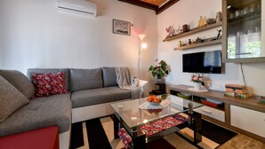 Una sala de estar con un sofá gris, una mesa de centro de vidrio, una silla roja, un televisor, estantes con objetos decorativos y un techo de madera.