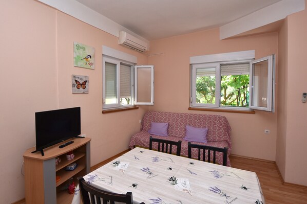 A small living room with a sofa, a TV on a stand, and two windows with blinds.