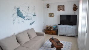 Un salon avec un canapé beige, une table basse en bois, un téléviseur et un mur orné d’œuvres d’art sur le thème de la mer.