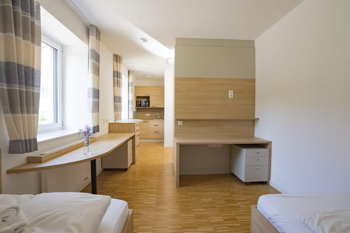 Una habitación de hotel moderna con suelo de madera, una cama, un escritorio y una kitchenette al fondo.