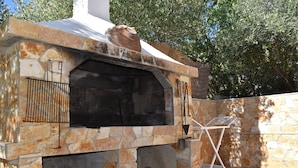An outdoor stone-built barbecue area with a chimney, a metal rack, and a wooden bench.