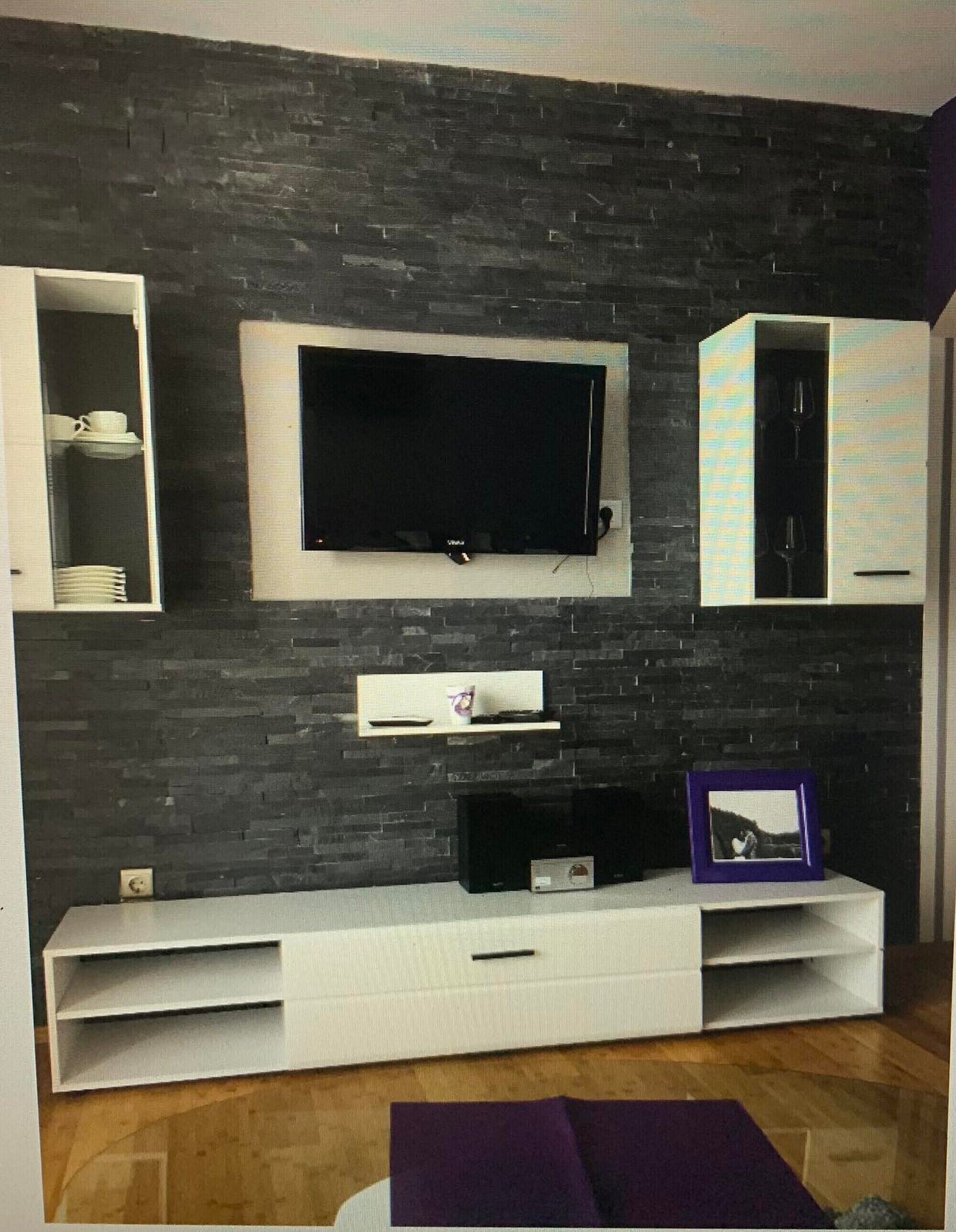 A modern living room with a flat-screen TV mounted on a dark brick wall, white built-in shelving units, and a wooden floor.