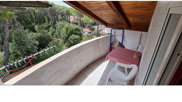 Ein Balkon mit rot-weiß kariertem Tisch, einem weißen Stuhl und Blick auf ein bewaldetes Gebiet.