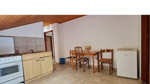 Eine kleine Küche mit Herd, Backofen und einem kleinen Kühlschrank. Es gibt einen Esstisch mit Stühlen und eine blaue Mülltonne.