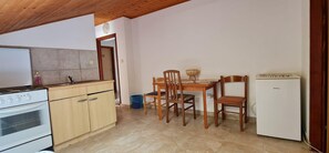 Eine kleine Küche mit Herd, Backofen und einem kleinen Kühlschrank. Es gibt einen Esstisch mit Stühlen und eine blaue Mülltonne.