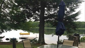 A glass-top table with four chairs and a blue umbrella, set on a grassy area near a lake. - Belgrade Lakes McGrath Pond (Oakland)