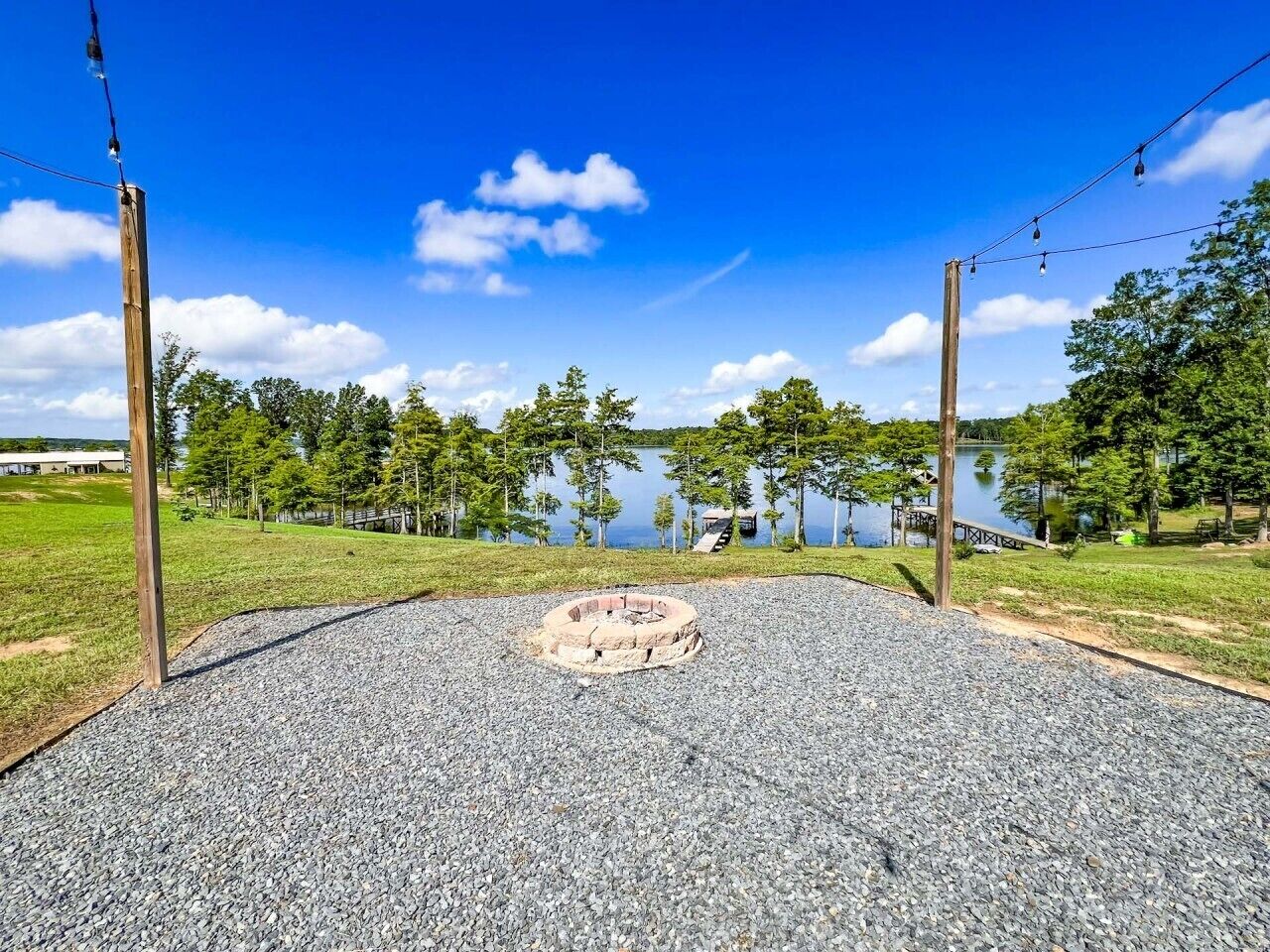 Un espace recouvert de gravier avec un foyer circulaire, entouré d’un champ herbeux et d’arbres, avec un lac et un quai en arrière-plan.