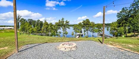Un área de grava con una fogata circular, rodeada por un campo cubierto de césped y árboles, con un lago y un muelle al fondo.
