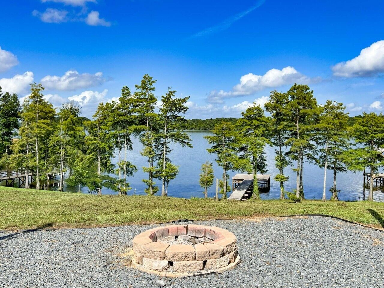 A lakeside setting with a fire pit, trees, and a dock.