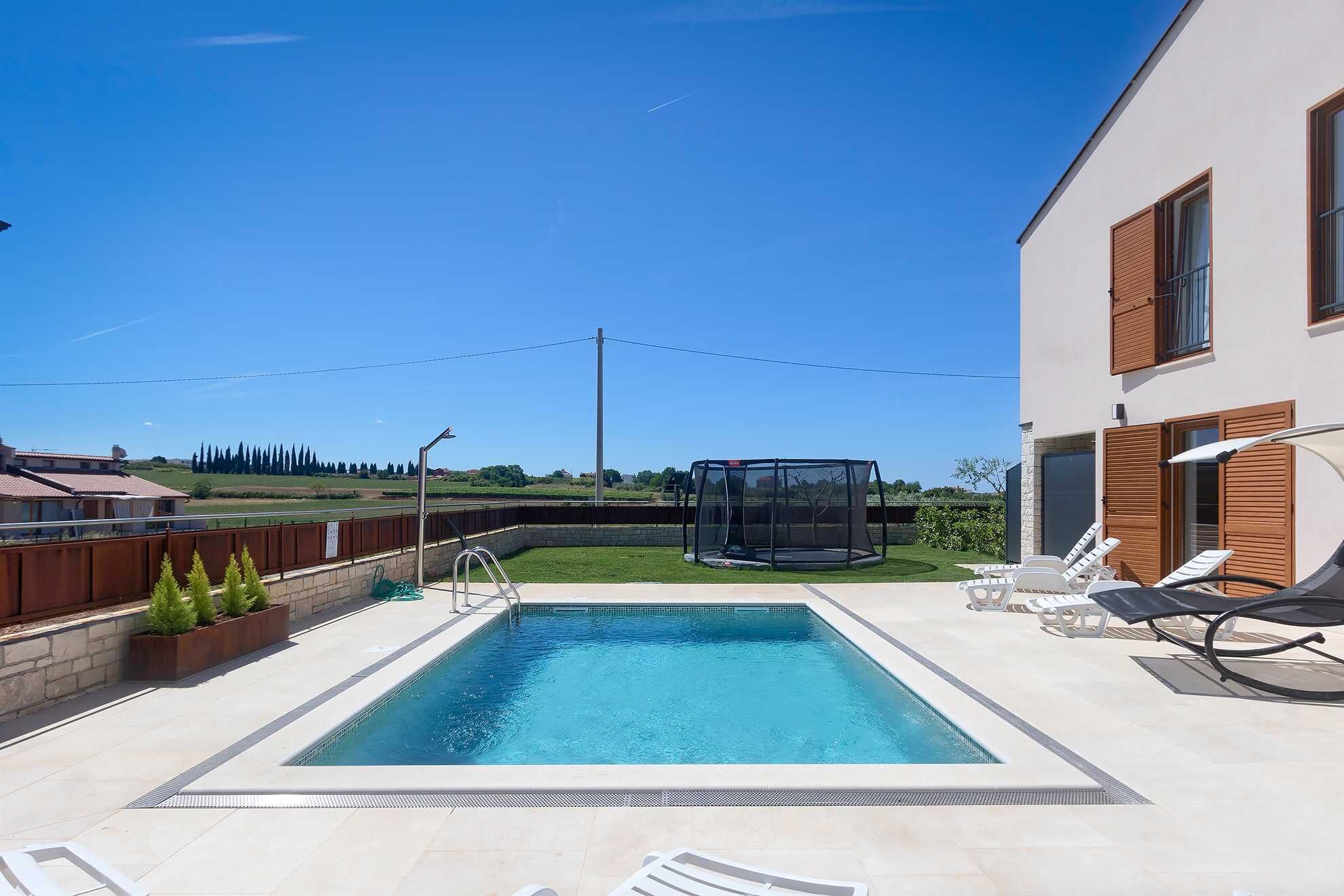 Una piscina rectangular con trampolín, rodeada de sillas de descanso y una zona de estar cubierta.
