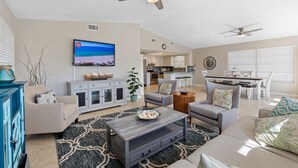 Een ruime woonkamer met een grote flatscreen-tv, plafondventilatoren en een keuken in de achtergrond.