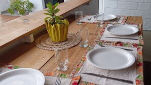 A wooden dining table set with white plates, cutlery, and clear glasses, with a potted plant and a shelf with books in the background. - Pousada Estresse Zero (Barreirinhas)