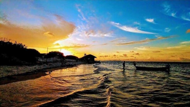 A sunset over a calm body of water with a boat and buildings in the background. - Pousada Estresse Zero (Barreirinhas)