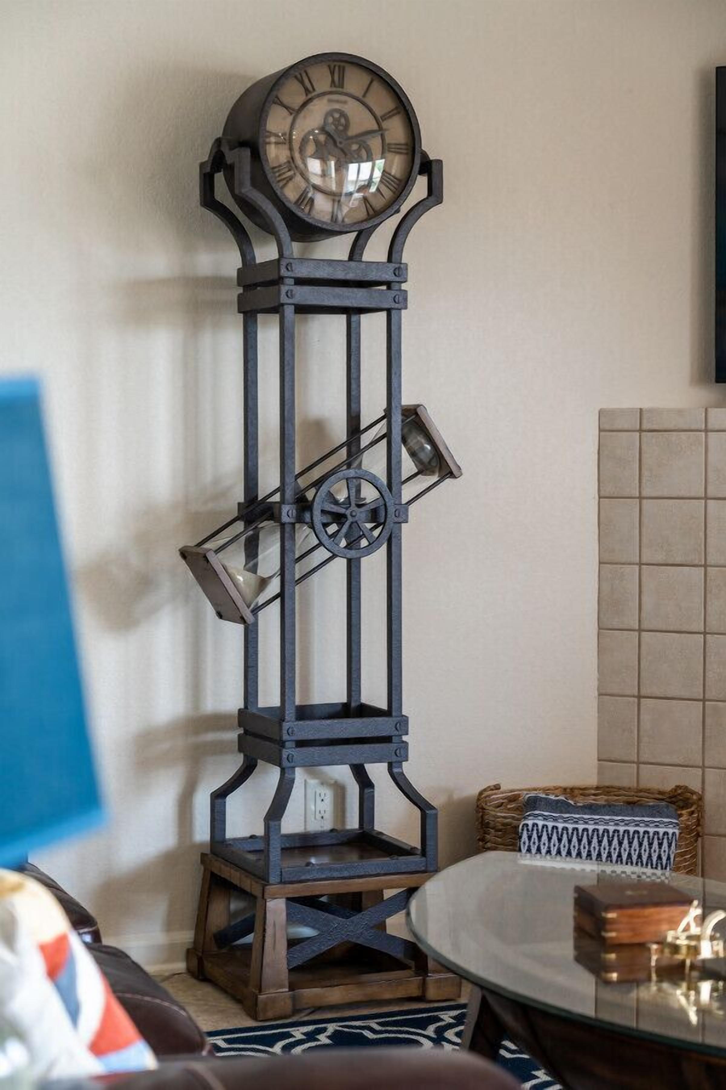 Un reloj de pie alto y ornamentado con números romanos, un péndulo y un peso sobre un soporte, colocado en una habitación con pared de azulejos y una mesa redonda.