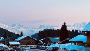 Un estacionamiento cubierto de nieve con varios edificios de madera, un paisaje de montañas al fondo y cielo despejado.