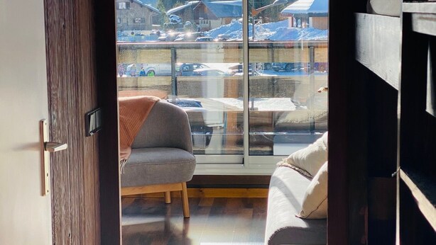 Una habitación moderna con un amplio ventanal corredero que da a un balcón. La habitación tiene un sofá, una mesita y una estantería.