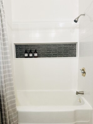 A modern bathroom with a white bathtub, a showerhead, and a shelf with toiletries. - Luxury Historic Home - Stellar Location (Milwaukee)