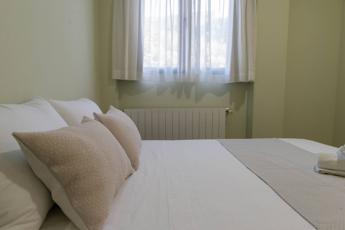 A neatly made bed with white linens and beige pillows in a room with a radiator and a window with sheer curtains.