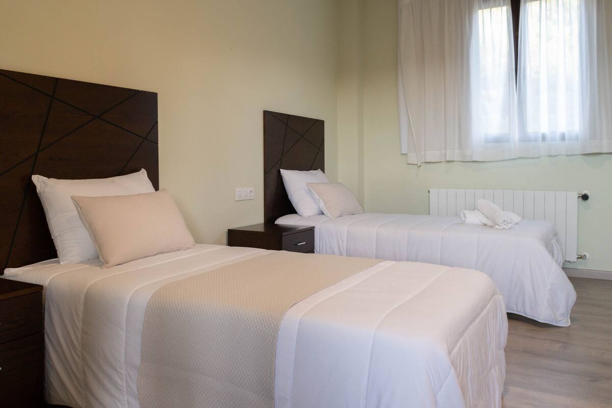 A hotel room with two beds, each with a wooden headboard, white bedding, and a window with sheer curtains.