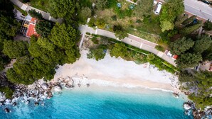 Luftaufnahme eines Strandes mit sandigem Ufer, klarem, blauem Wasser und umgebender Bepflanzung.