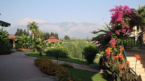 A landscaped garden with pathways, a building with a red roof, and mountains in the background.