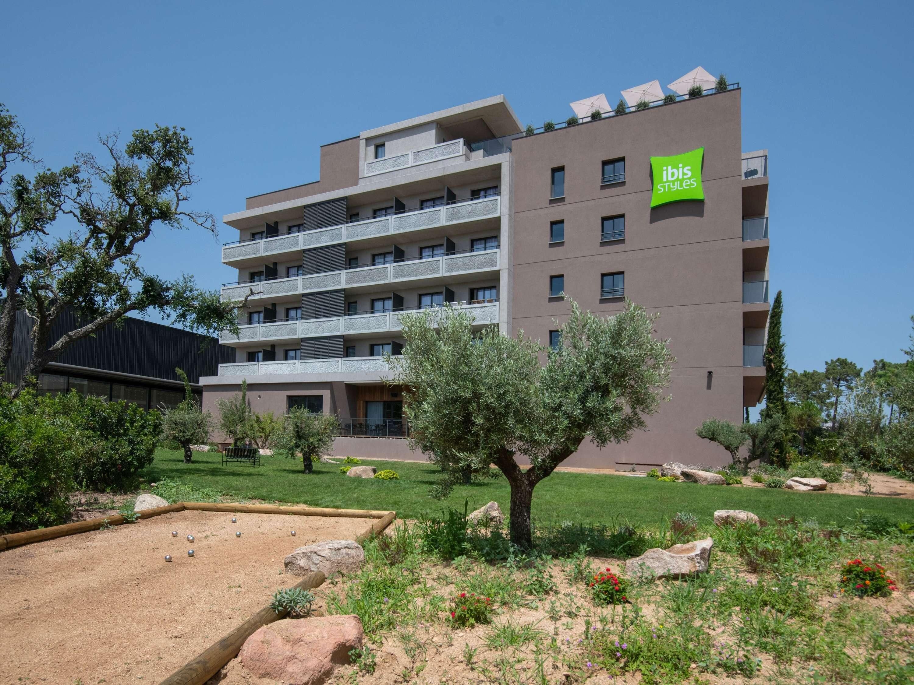 Um hotel com vários andares, varandas, um jardim com pedras e plantas, e uma placa com a inscrição 'ibis Styles'.