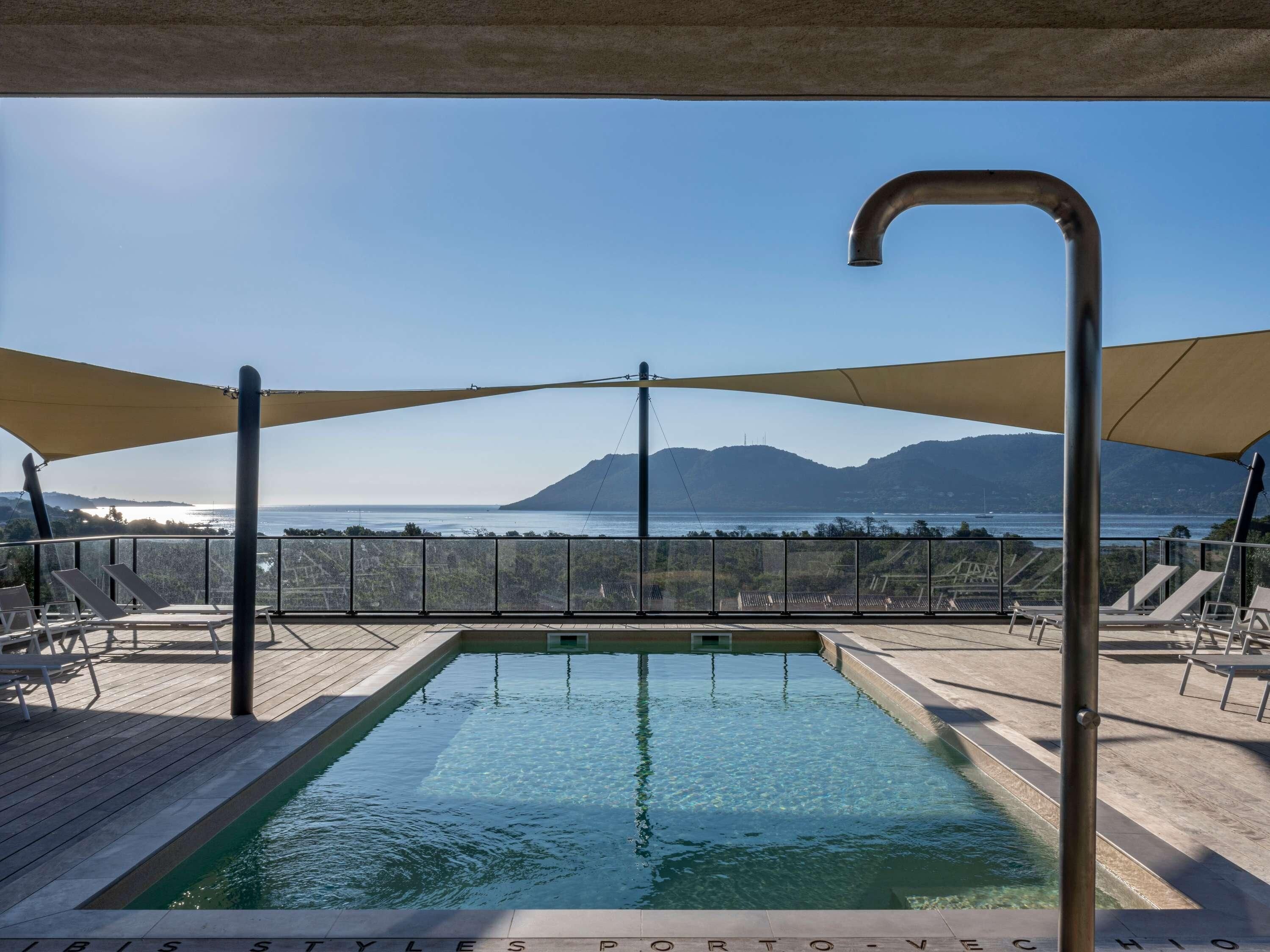 An outdoor swimming pool with a view of the sea and mountains, surrounded by lounge chairs and shaded by large canopies.