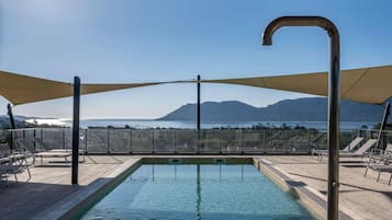 An outdoor swimming pool with a view of the sea and mountains, surrounded by lounge chairs and shaded by large canopies.
