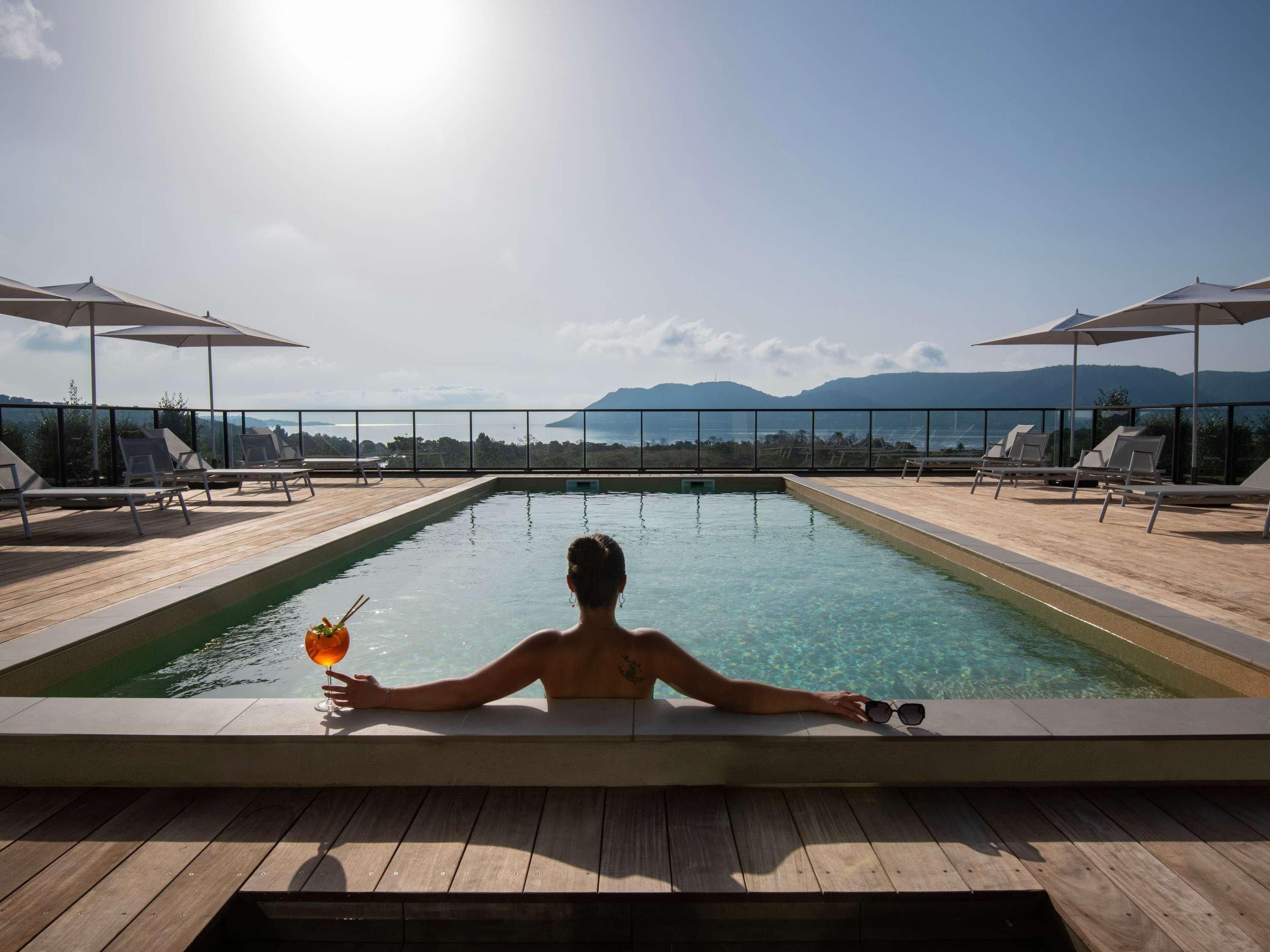 Una persona relajándose en una piscina infinita iluminada por el sol, con agua cristalina, rodeada de sillas de madera y sombrillas, con vistas a un paisaje montañoso.