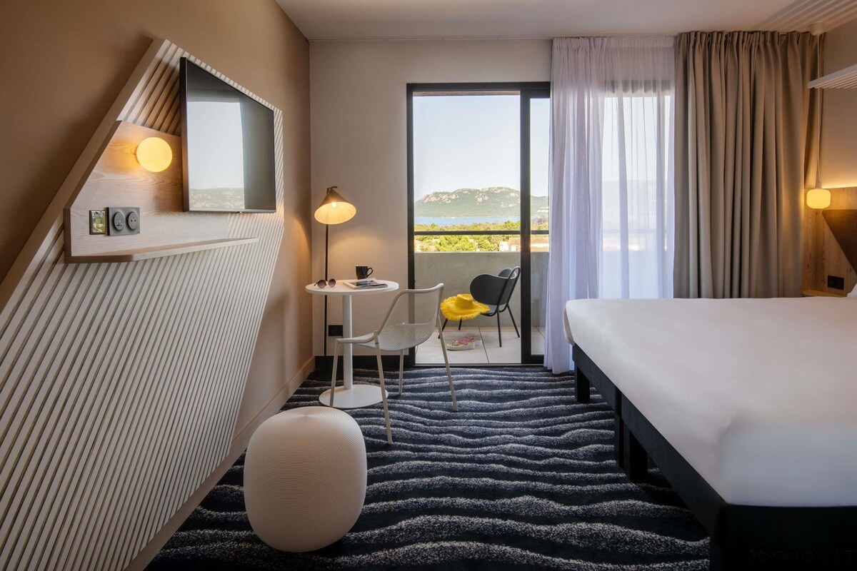 A hotel room with a bed, a small table with a chair, a round ottoman, a mirror, and a view of the sea through a balcony.