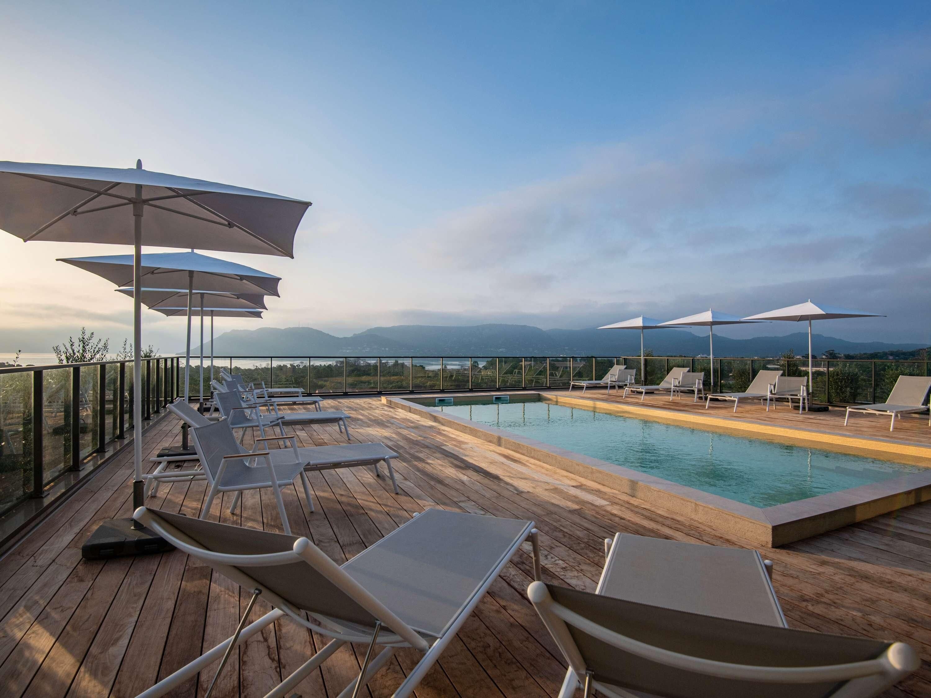 A rooftop pool area with lounge chairs and umbrellas, overlooking a mountainous landscape.