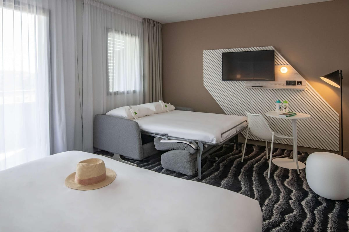 A hotel room with a bed, a desk, a chair, a TV, and a hat on the bed.
