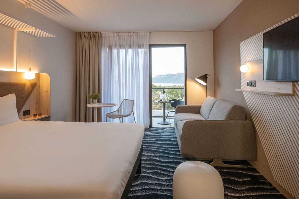Habitación de hotel con cama, sofá, una mesita con silla, televisor montado en la pared y vista al exterior.