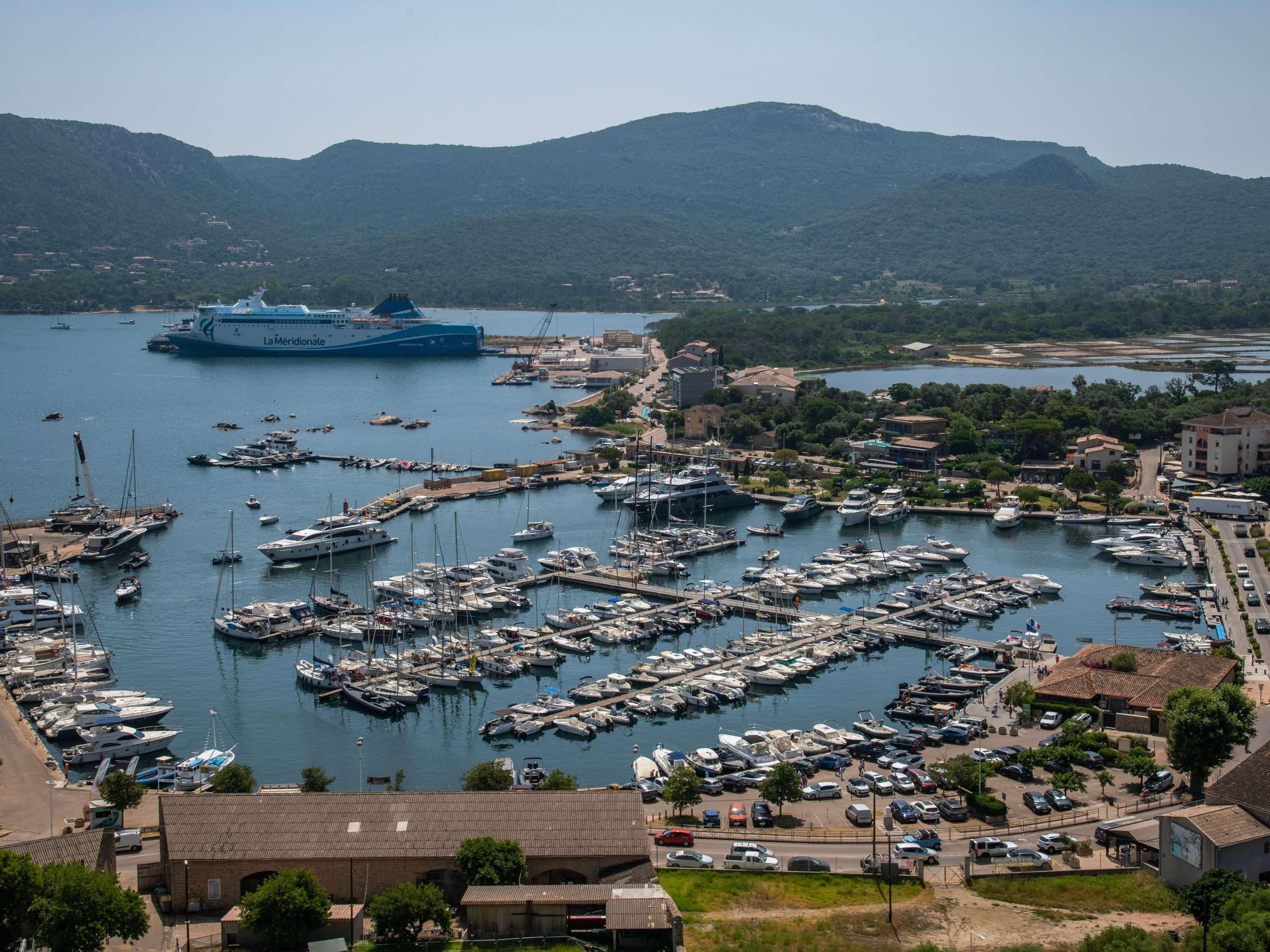 Un port de plaisance où sont amarrés de nombreux bateaux, un grand navire de croisière en arrière-plan et une ville avec des bâtiments et des véhicules.
