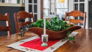 Une table à manger avec un set de table rouge, un porte-bougie blanc orné d’un motif de sapin de Noël et un bol rempli de plantes vertes.