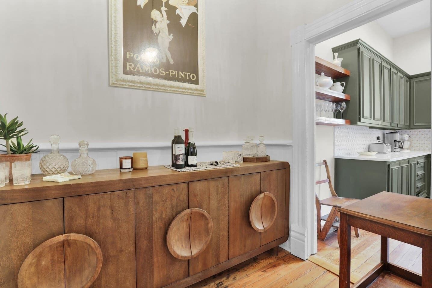 A wooden dining table with a chair, a kitchen area with green cabinets, and a wall-mounted wine bottle holder with two bottles.