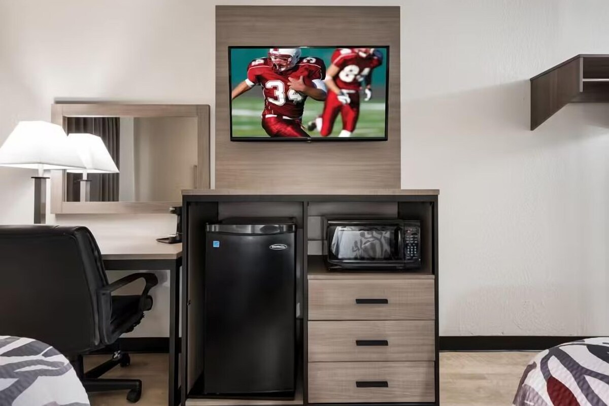 A compact hotel room with a desk, microwave, refrigerator, and a TV displaying a sports game.
