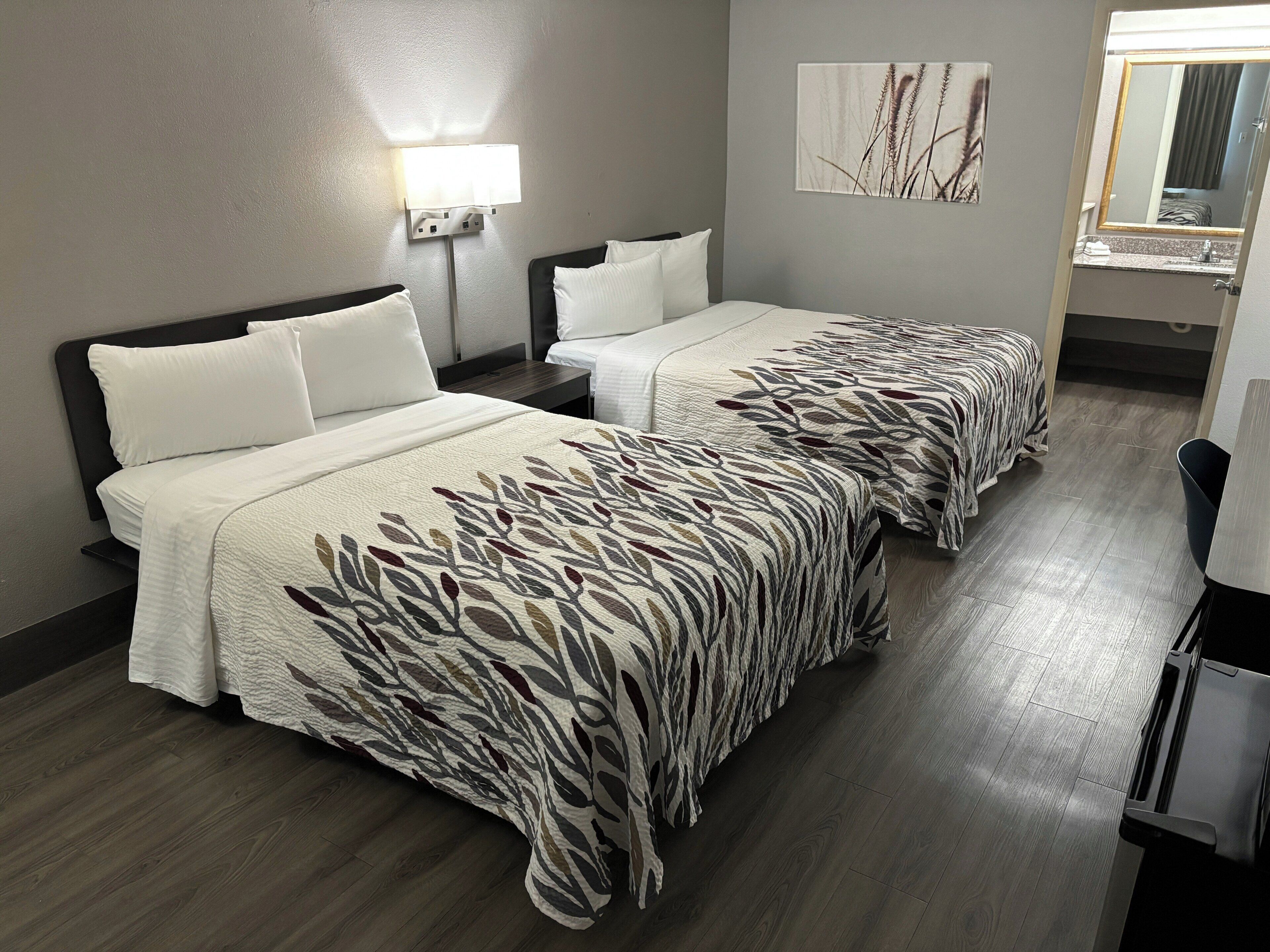 A hotel room with two beds, a nightstand, a lamp, a mirror, and a painting on the wall.