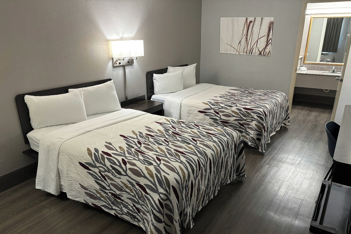 A hotel room with two beds, a nightstand, a lamp, a mirror, and a painting on the wall.