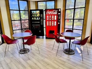 A room with two round tables and red chairs, a Coca-Cola vending machine, and large windows with a view of greenery. - Red Roof Inn Birmingham (Birmingham)