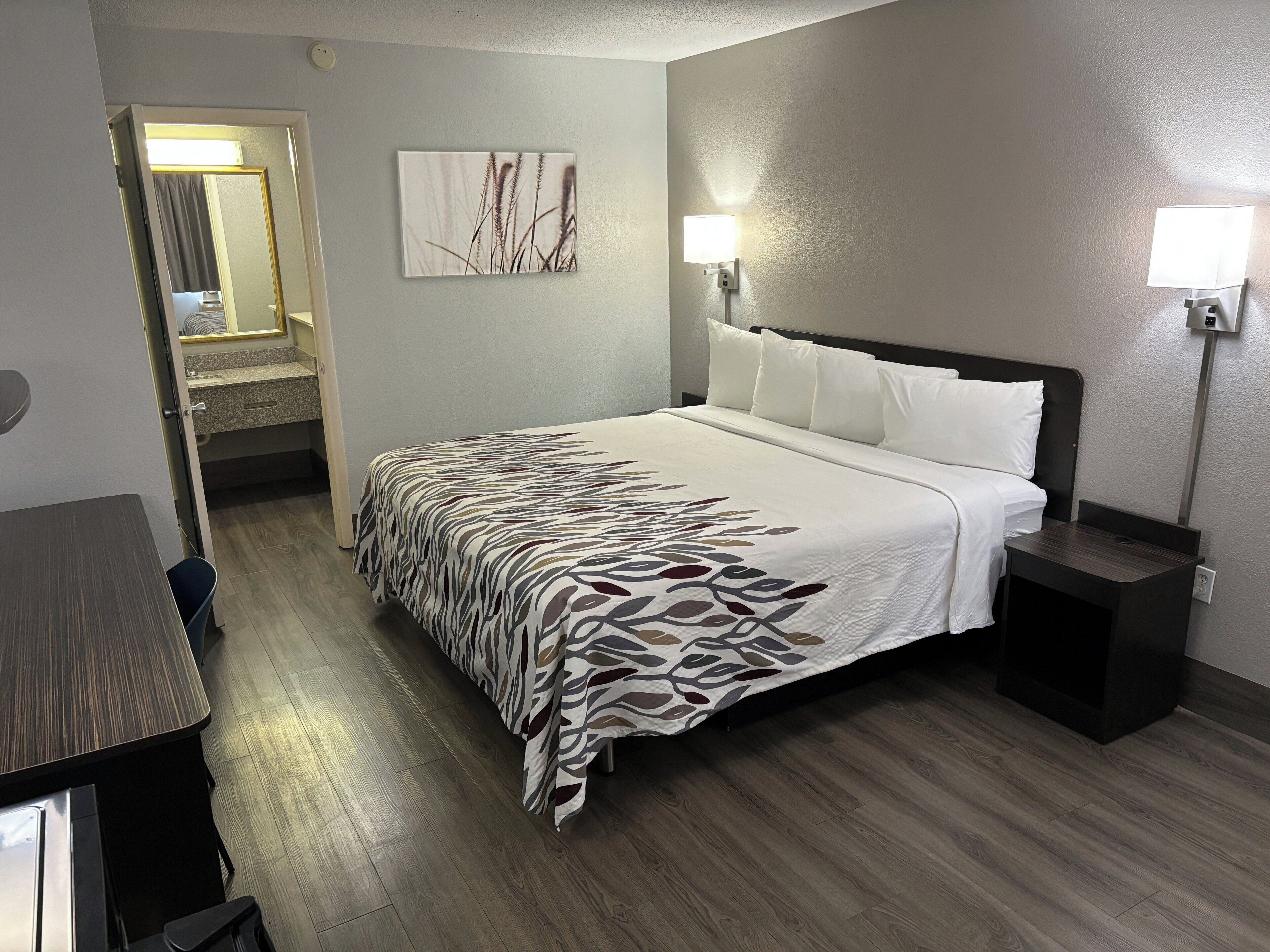 A hotel room with a bed, bedside tables, a nightstand, a wall-mounted lamp, and a door leading to a bathroom.