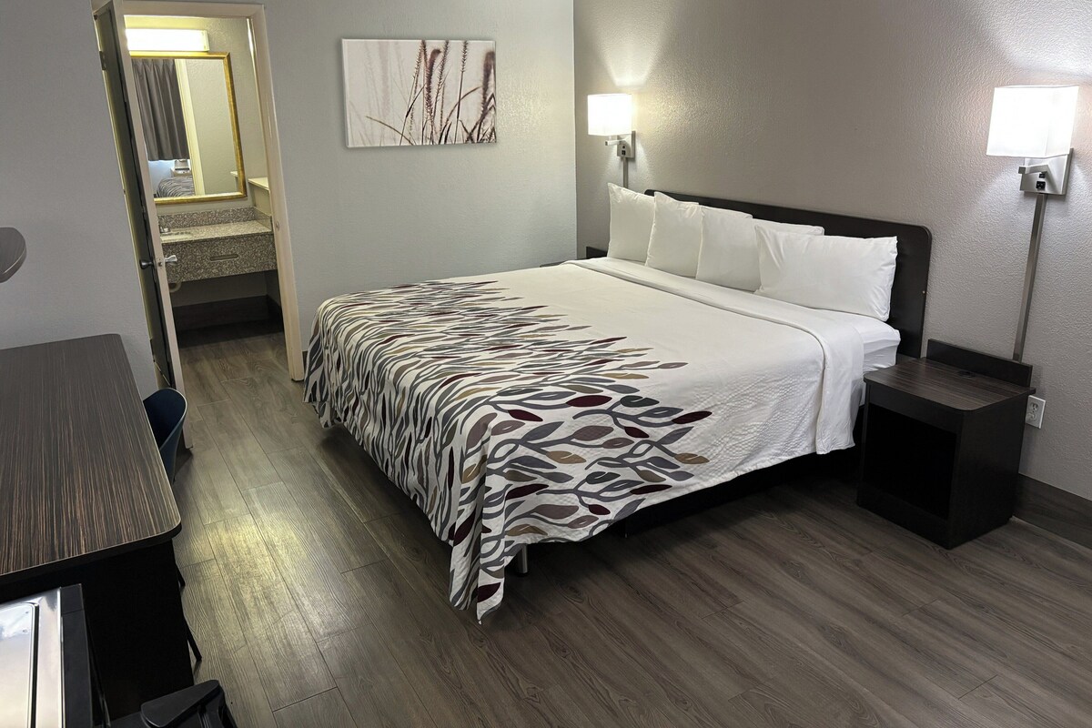 A hotel room with a bed, bedside tables, a nightstand, a wall-mounted lamp, and a door leading to a bathroom.