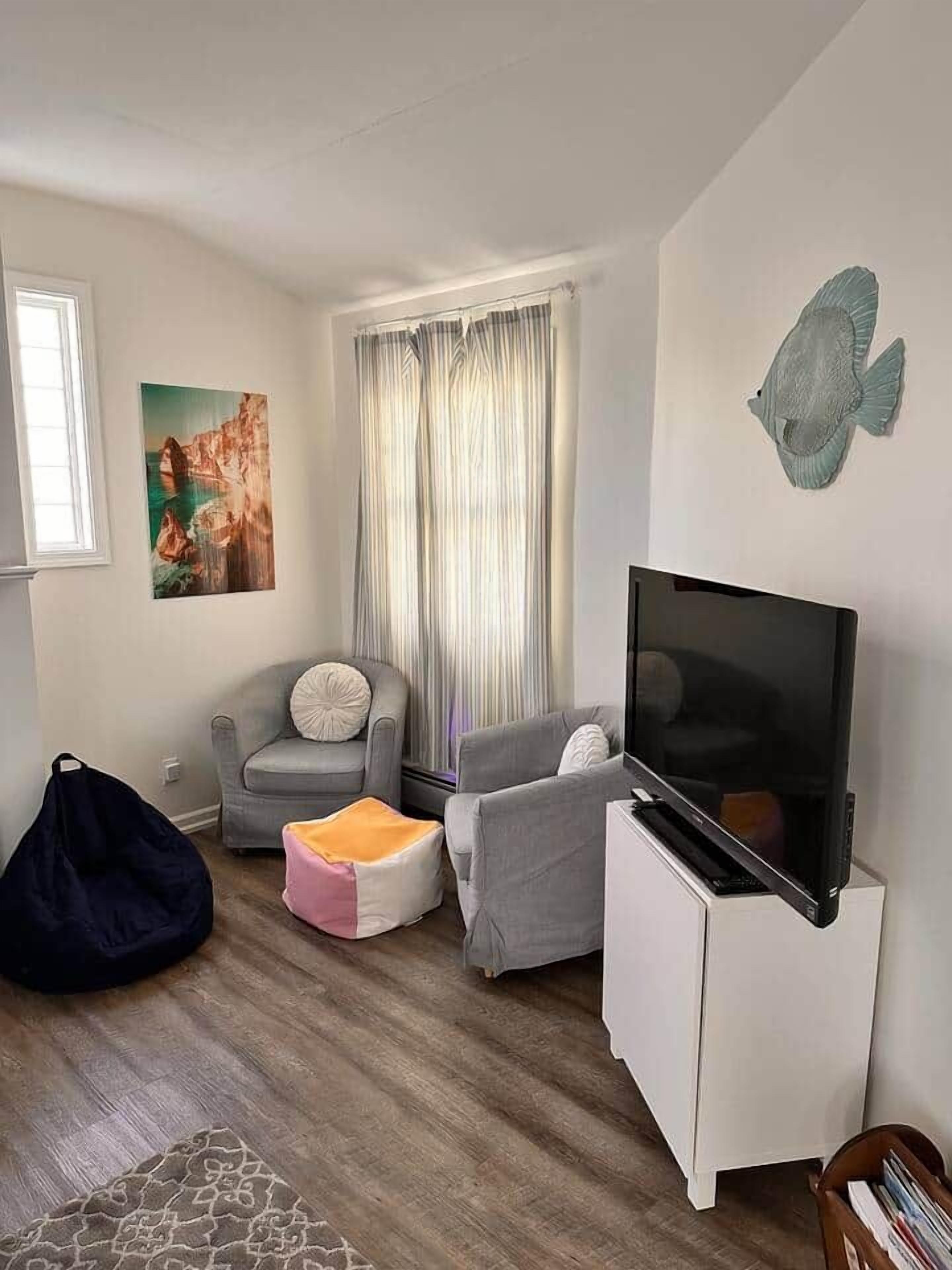 Een woonkamer met een flatscreen televisie, een grijze bank, een witte kast, een loungestoel en een vloerkleed met dessin.