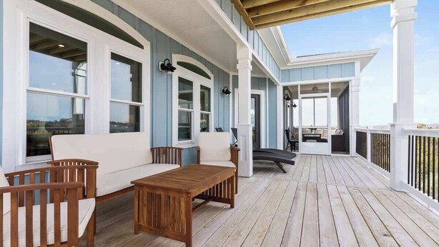 A covered outdoor deck with wooden furniture, large windows, and a view of the landscape. - Ocean Twelve (Ocean Isle Beach)