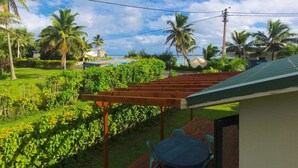 A wooden deck with a table and chairs, surrounded by lush greenery and palm trees, overlooking a beach and ocean. - MM Batch South Side (Avavaaroa)