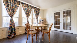 Comedor con una mesa redonda y seis sillas de mimbre, ventanas grandes con cortinas florales y una puerta blanca con paneles de vidrio.