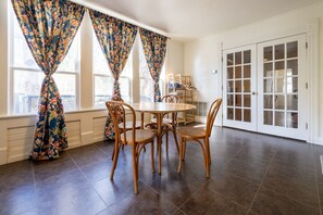 A dining area with a round table and six wicker chairs, large windows with floral curtains, and a white door with glass panels.