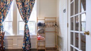 A room with a window, a wooden shelf with hanging clothes, and a door with a brass doorknob. - The Downtown Abbey - The Drawing Room (San Marcos)