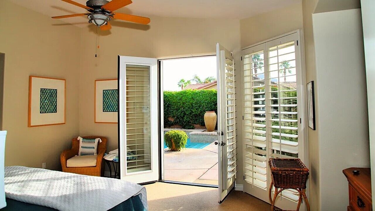 Une chambre avec un lit, une chaise, un ventilateur de plafond et une vue sur une piscine, visible à travers de grandes portes-fenêtres ouvertes.