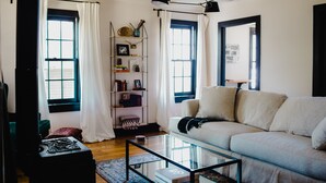 A living room with a sofa, a glass coffee table, a bookshelf, and a window with curtains. - The Starling at Pond Eddy, Cottage (Pond Eddy)